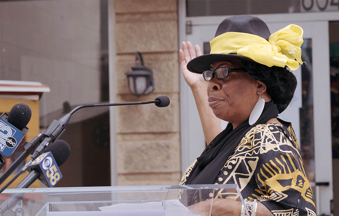 A woman speaking at the podium points up to the mural.
