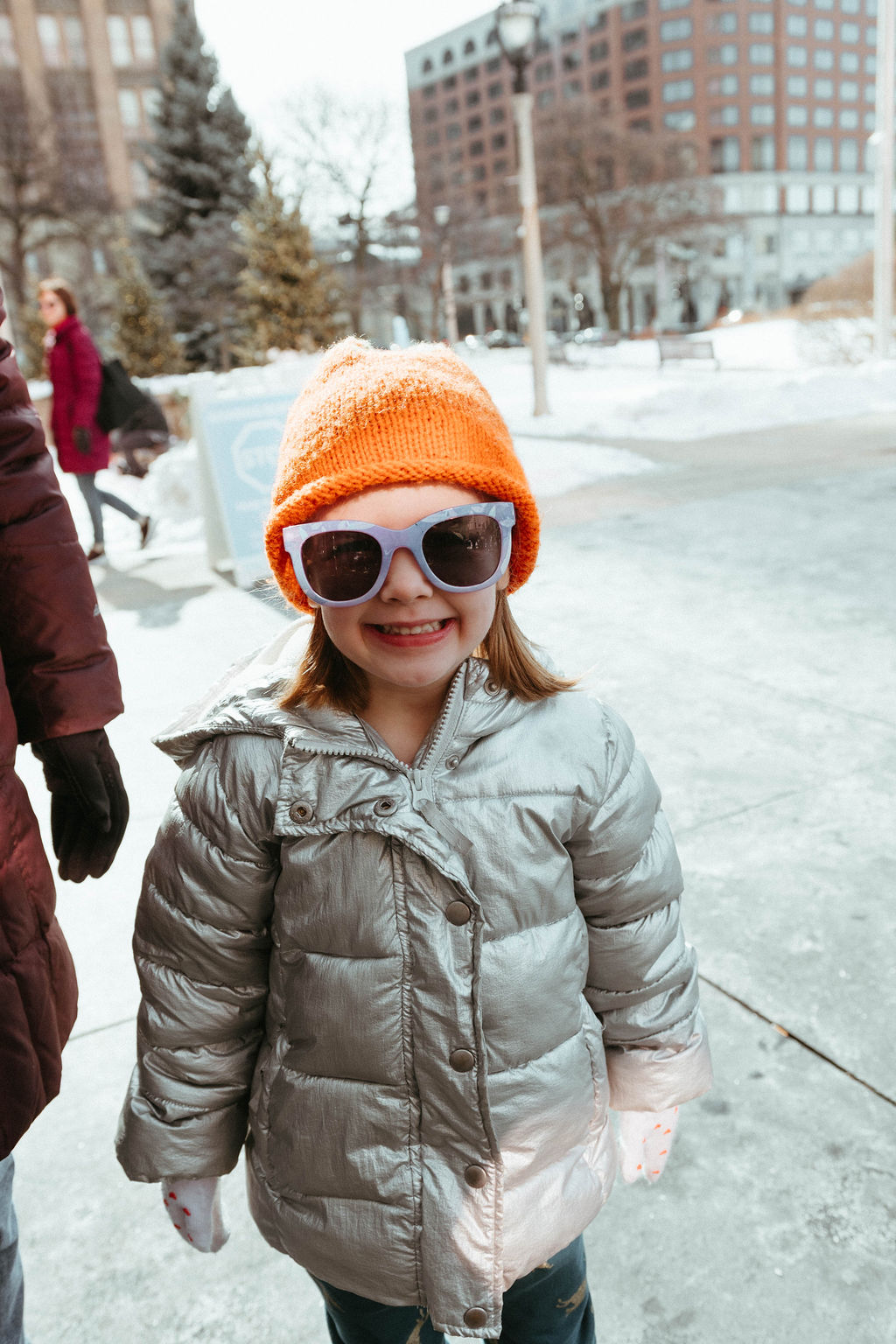 A little girl wears sunglasses and a bright orange hat on a snowy day.